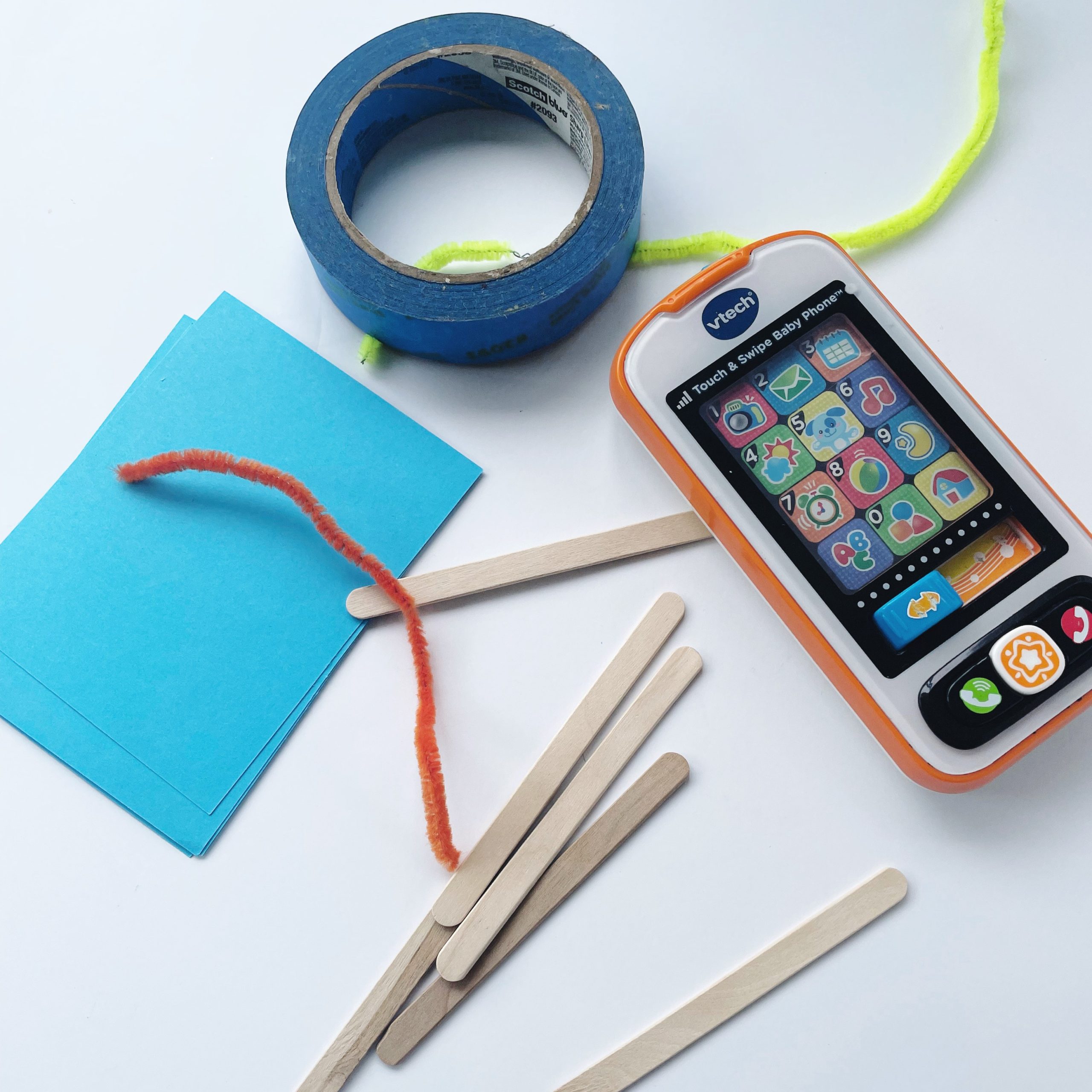 Vtech phone device, blue tape, popsicle sticks, blue paper, and a yellow pipe cleaner on a table as materials for an engineering design project as part of a what is engineering lesson